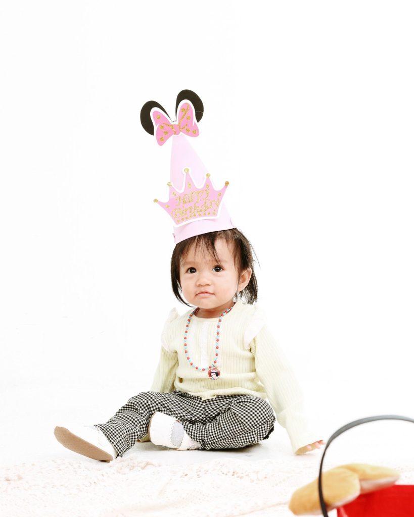 Baby in a pink party hat sitting on lace, first birthday photoshoot at The Cade & Co studio Cyberjaya
